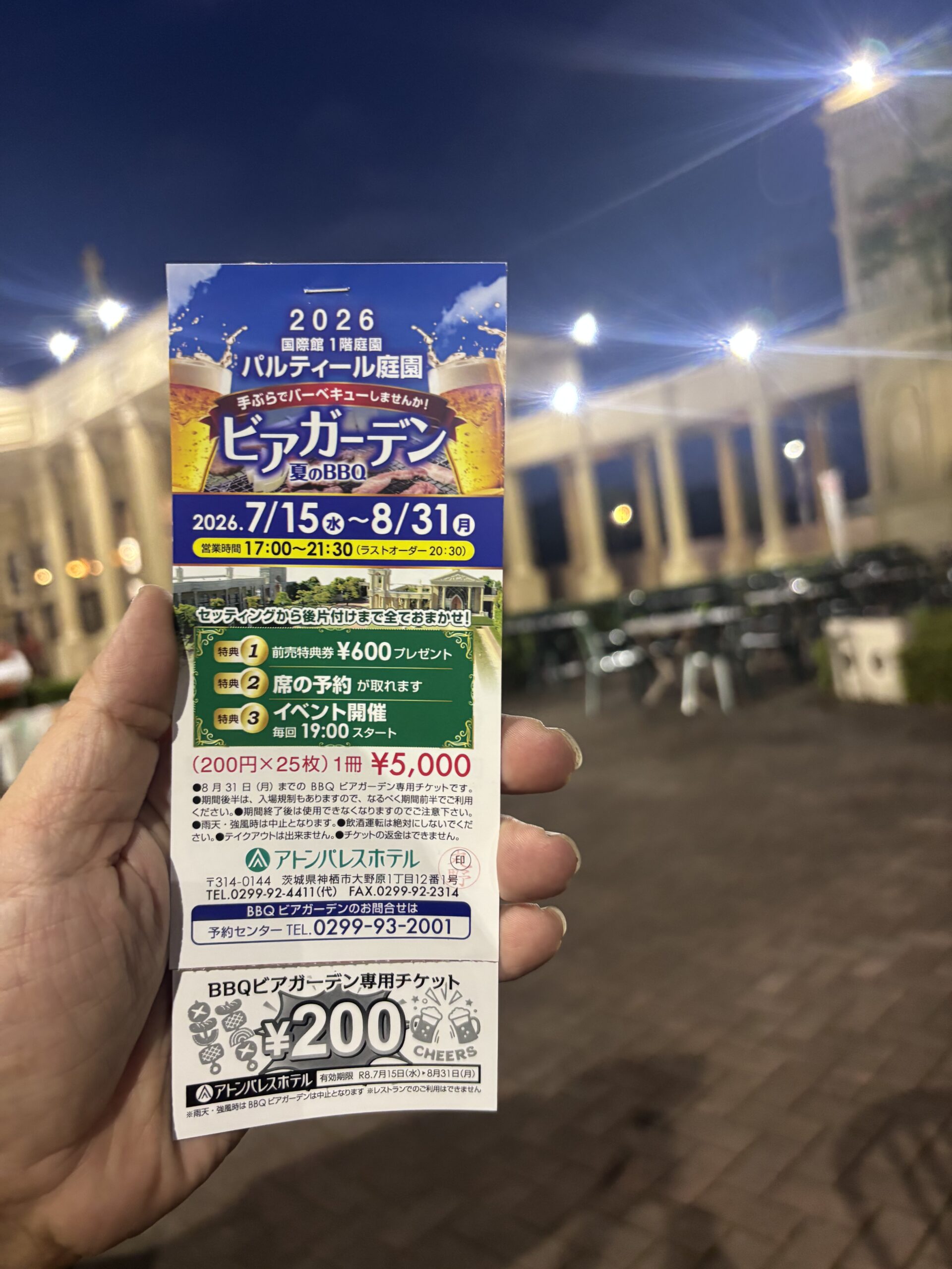アトンパレスホテル国際館1階庭園「パルティール庭園」のビアガーデン会場を背景に、手に持ったBBQビアガーデン専用チケットの冊子